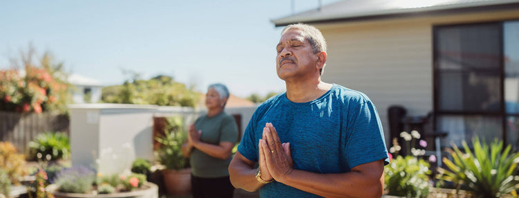 Man wearing a blue shirt meditating outside a house with plants.