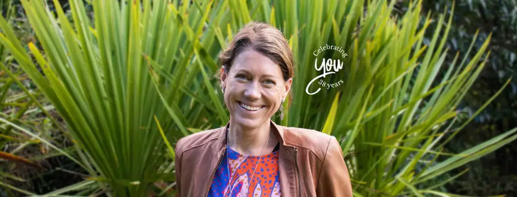 A smiling woman with short brown hair stands outdoors in front of large green palm-like leaves, wearing a brown jacket.