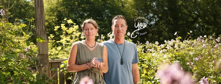 A man and a woman standing together in a sunlit garden with green foliage and flowers in the background.