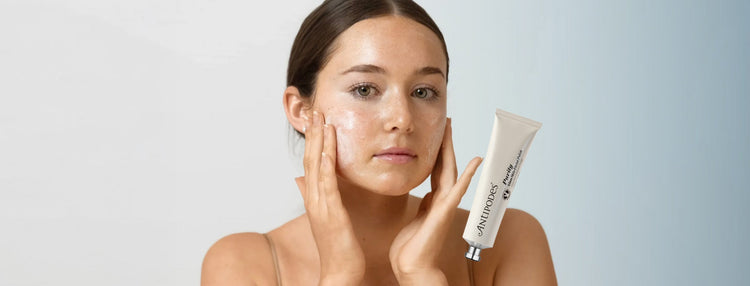Model applying Antipodes Purity Glass Skin Facial Polish to her cheeks, with a pale blue gradient background. Image overlay of the facial polish is next to the model's face.