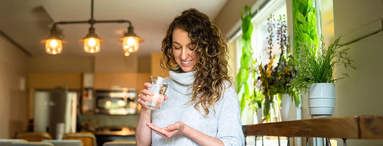 Woman with curly hair wearing a grey turtleneck while holding a glass of water with a supplement in the other hand, with a kitchen backdrop and indoor plants as decorative elements
