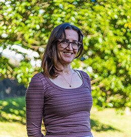 Woman wearing glasses and a purple shirt standing outdoors with greenery in the background