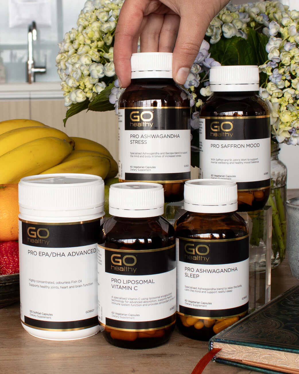 Collection of GO Healthy supplement bottles on a kitchen counter with flowers and bananas in the background.