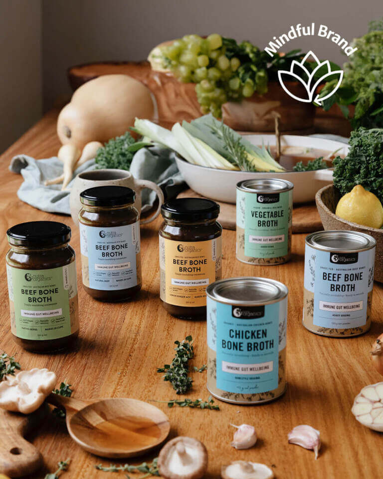Assorted bone broth products on a wooden table with fresh vegetables and herbs.