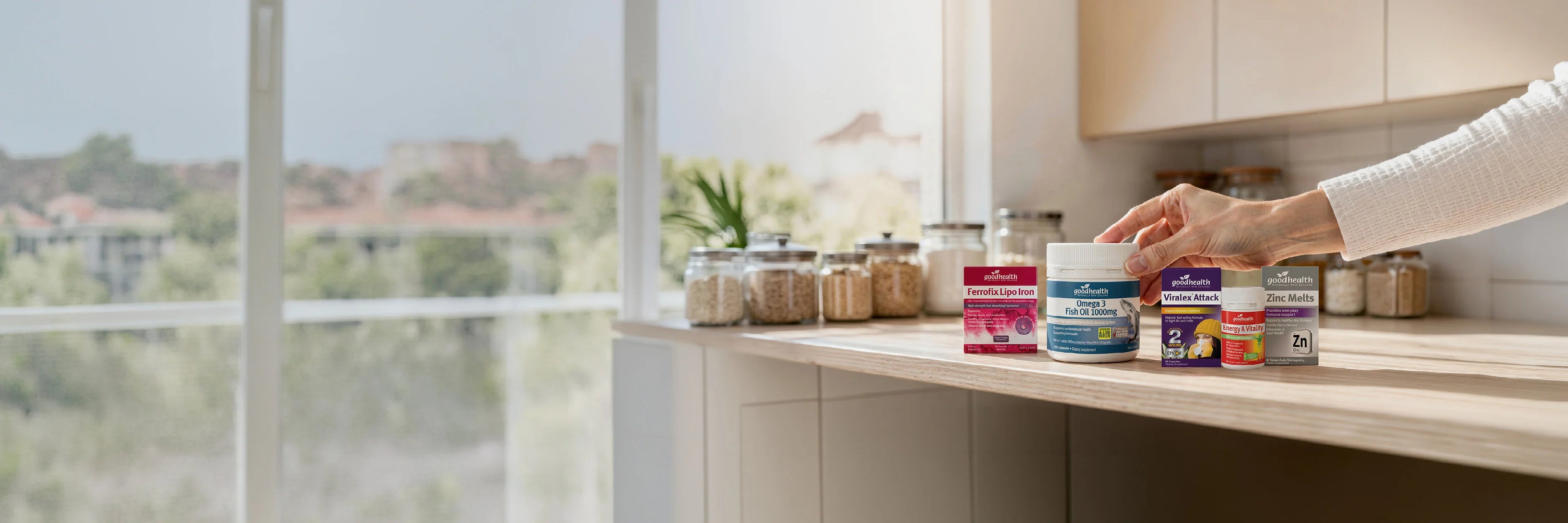 Person reaching for a jar of Good Health Fish Oil on a kitchen counter with a view of trees outside.