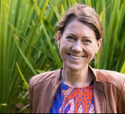A smiling woman with light brown hair stands outdoors in front of green palm leaves, wearing a brown leather jacket over a colorful patterned top. A white script watermark in the upper right reads 'Celebrating you 39 years'.