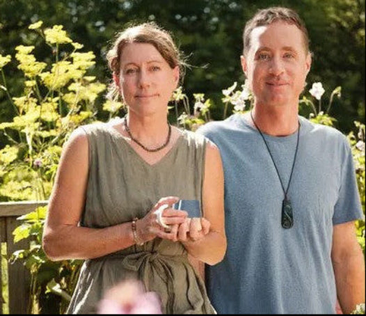 A man and a woman standing outdoors in a sunny garden with green foliage and flowers behind them. The woman wears an olive green sleeveless dress, and the man wears a light blue t-shirt.