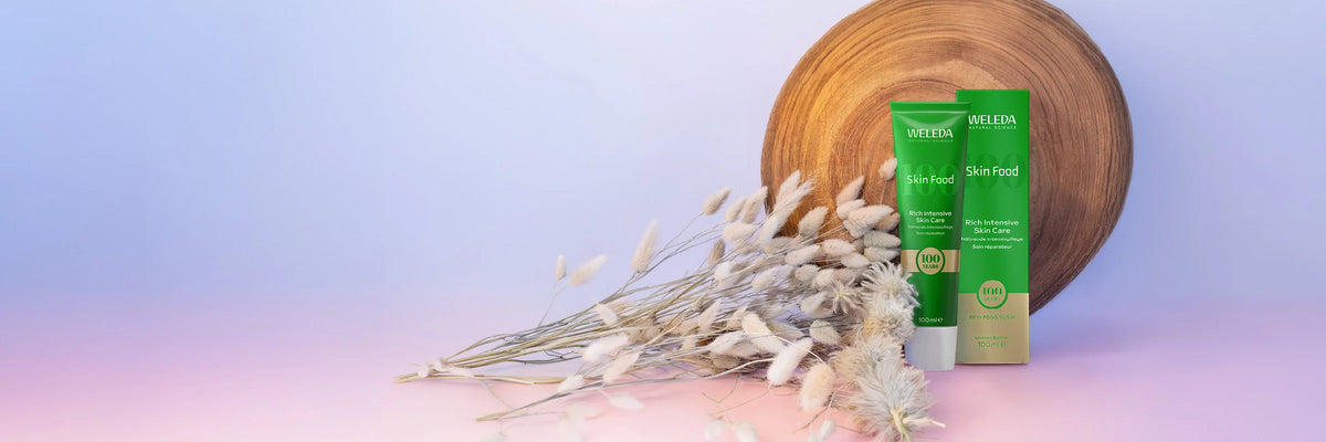 Weleda Skin Food celebrating 100 years packaging on a gradient background with a wooden tree trunk round and fluffy foliage.