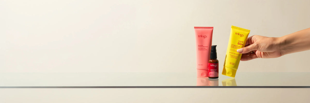 Trilogy Rosehip Cream Cleanser, SPF50+ Omega Boost Sheer Mineral Sunscreen & Instant Glow Rosehip Oil all positioned on a glass surface with a beige background with a hand holding the yellow sunscreen tube.