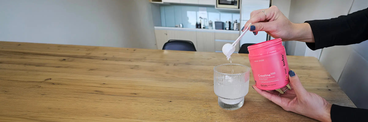 A person holding a pink tub of powder and pouring white powder into a glass of water using a scoop, set on a wooden table with a blurred kitchen background.