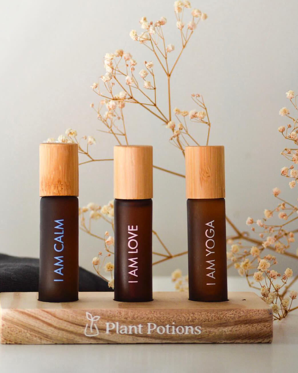 Three brown bottles with wooden caps labeled 'I AM CALM', 'I AM LOVE', and 'I AM YOGA' on a wooden block with 'Plant Potions' branding.