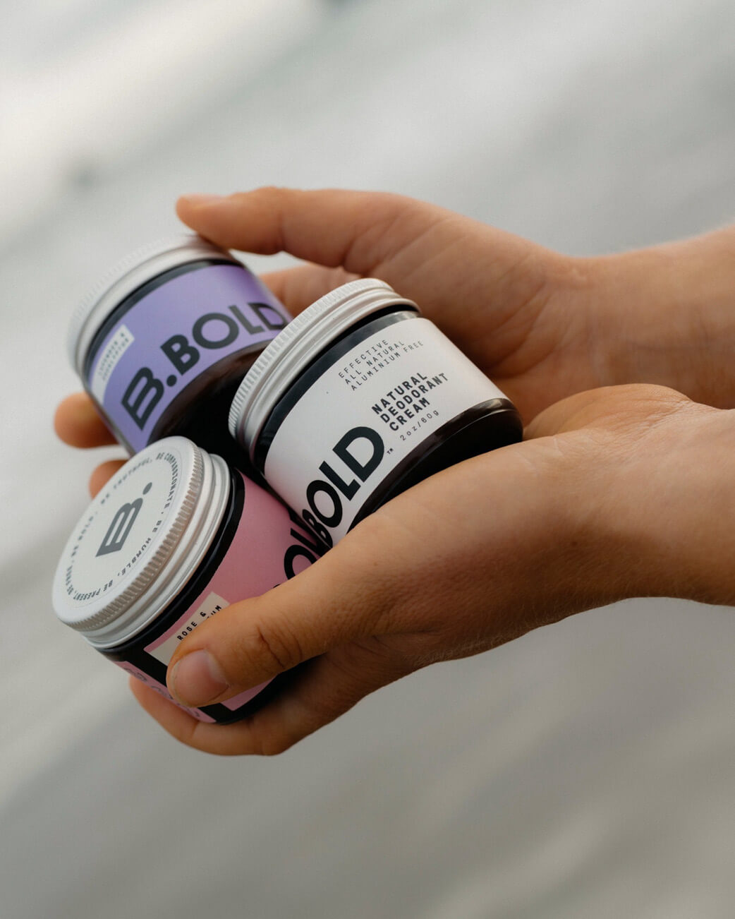 Hand holding three jars of B. Bold makeup products against a blurred background