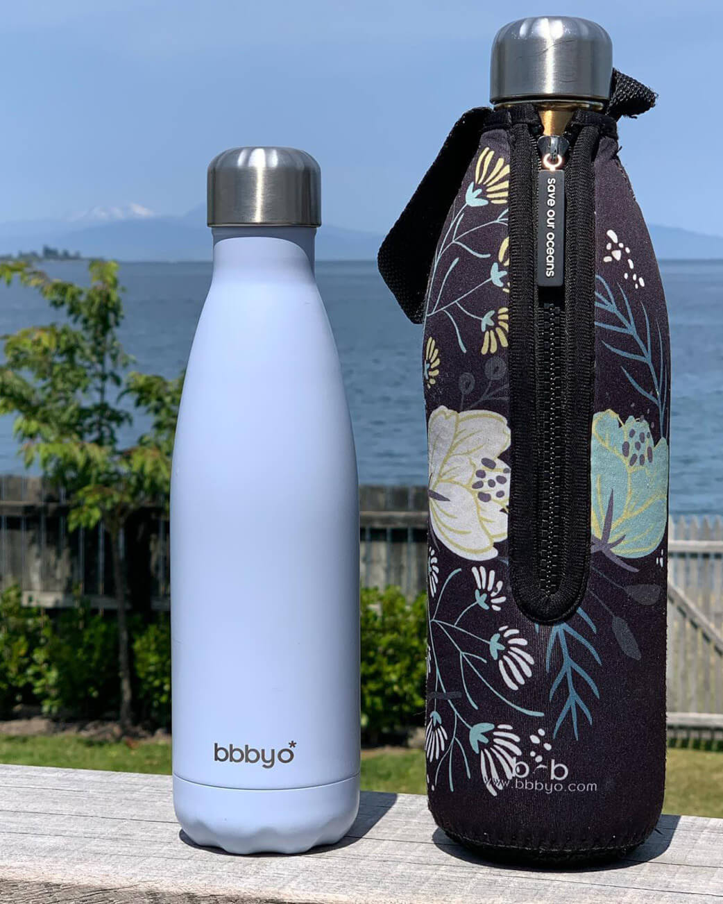 Two water bottles, one plain and one with a floral design, on a wooden surface with a scenic background.