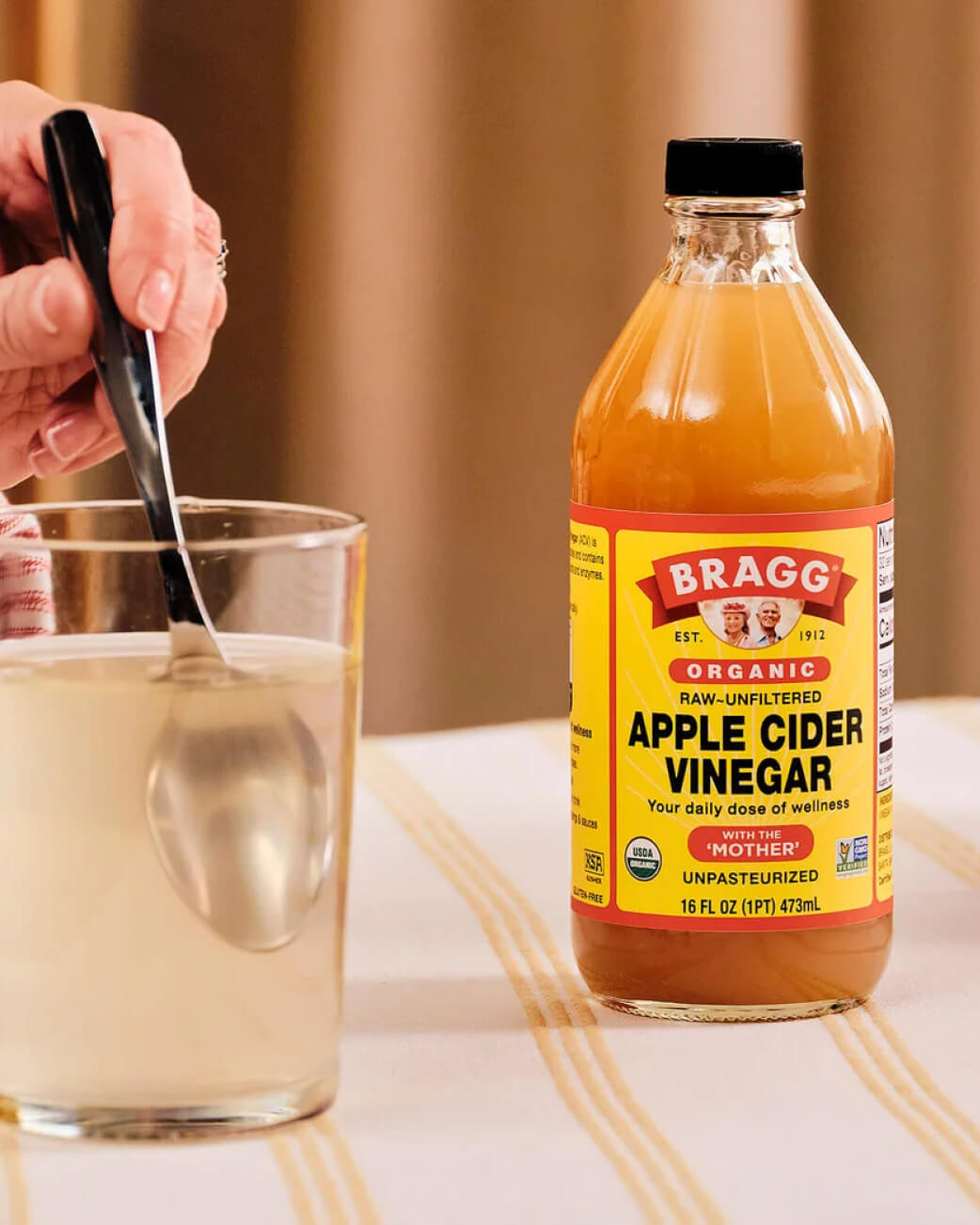 Bottle of Bragg Organic Apple Cider Vinegar next to a glass with a spoon on a table.