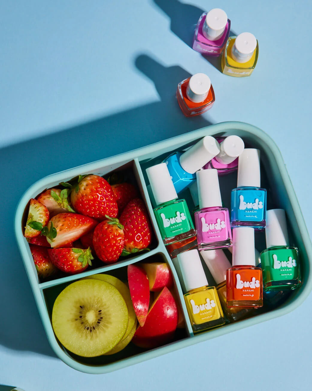 Bento box with strawberries, kiwi, and Buxom nail polish bottles on a blue background