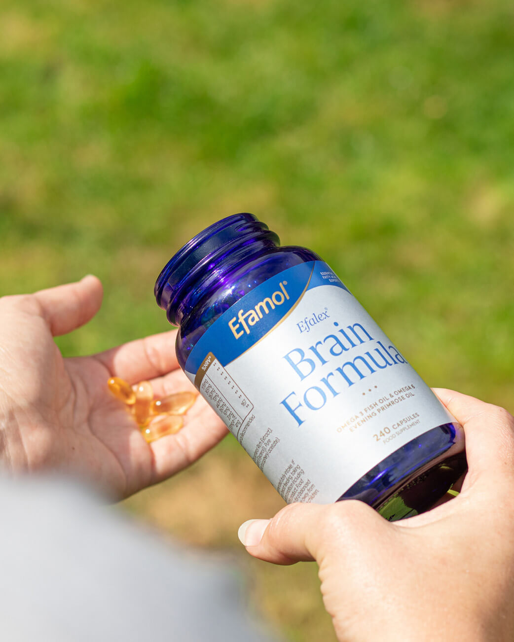 Person holding a bottle of Efamol Brain Formula supplement with a blurred green background.