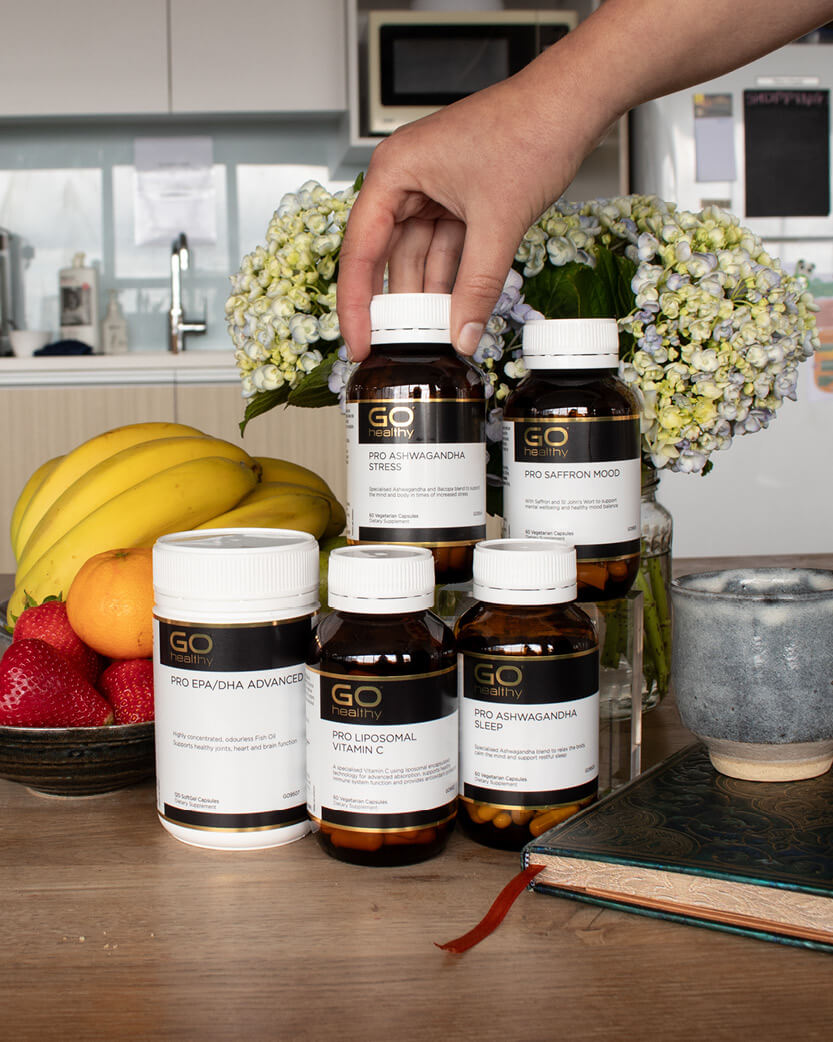 Hand picking a bottle of GO Nutrition supplements on a kitchen counter with fruits and flowers in the background.