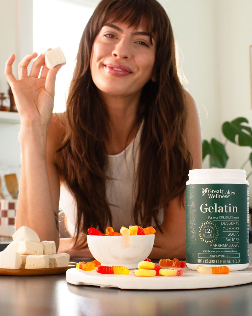 Woman holding a block of gelatin with a bowl of gummy bears and a container of Great Lakes Wellness Gelatin on a table.