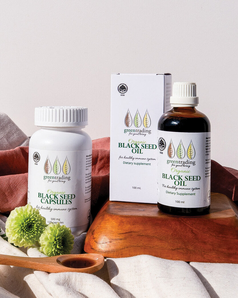 Black seed products including capsules, oil, and a box on a wooden surface with flowers.