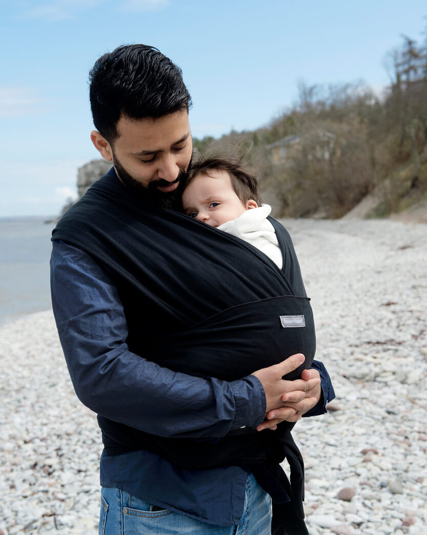Man holding a baby in a carrier on a pebbly beach with a scenic background.