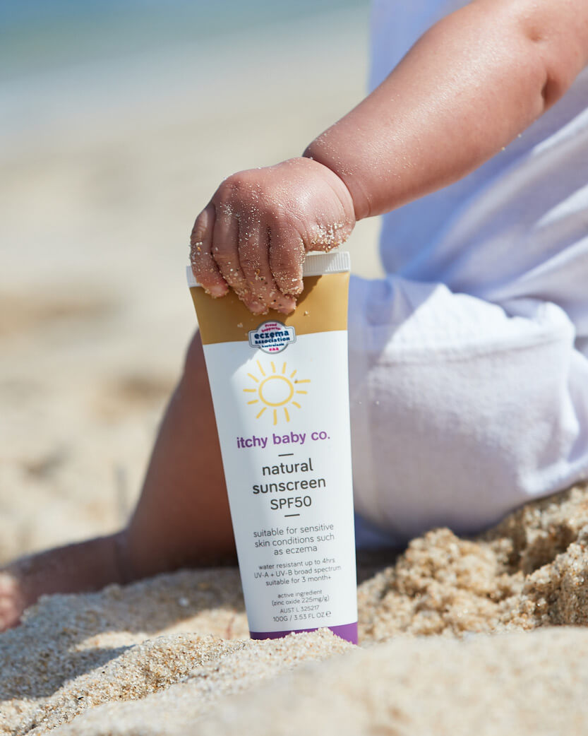 Child's hand holding an Itchy Baby Co. natural sunscreen SPF50 tube on sand.