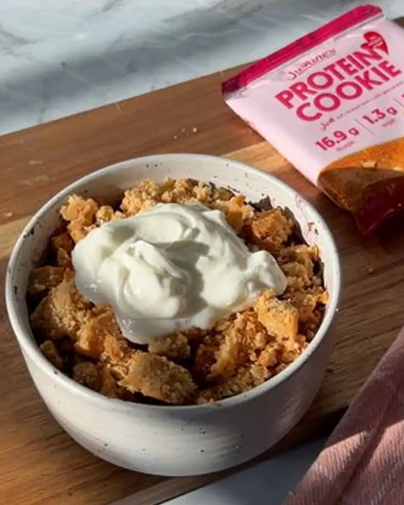 Bowl of crumble dessert with whipped cream next to a Protein Cookie package on a wooden surface.