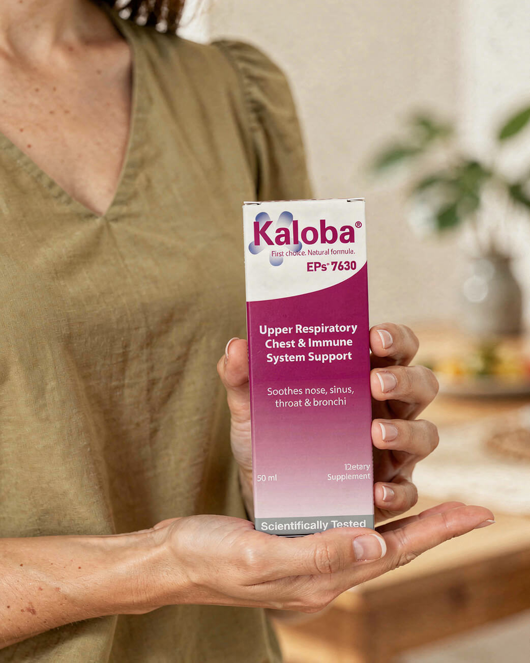 Person holding a Kaloba supplement box in a blurred indoor setting.