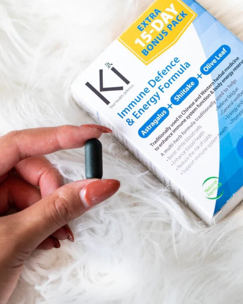Hand holding a supplement capsule with KI Immune Defence & Energy Formula packaging in the background.