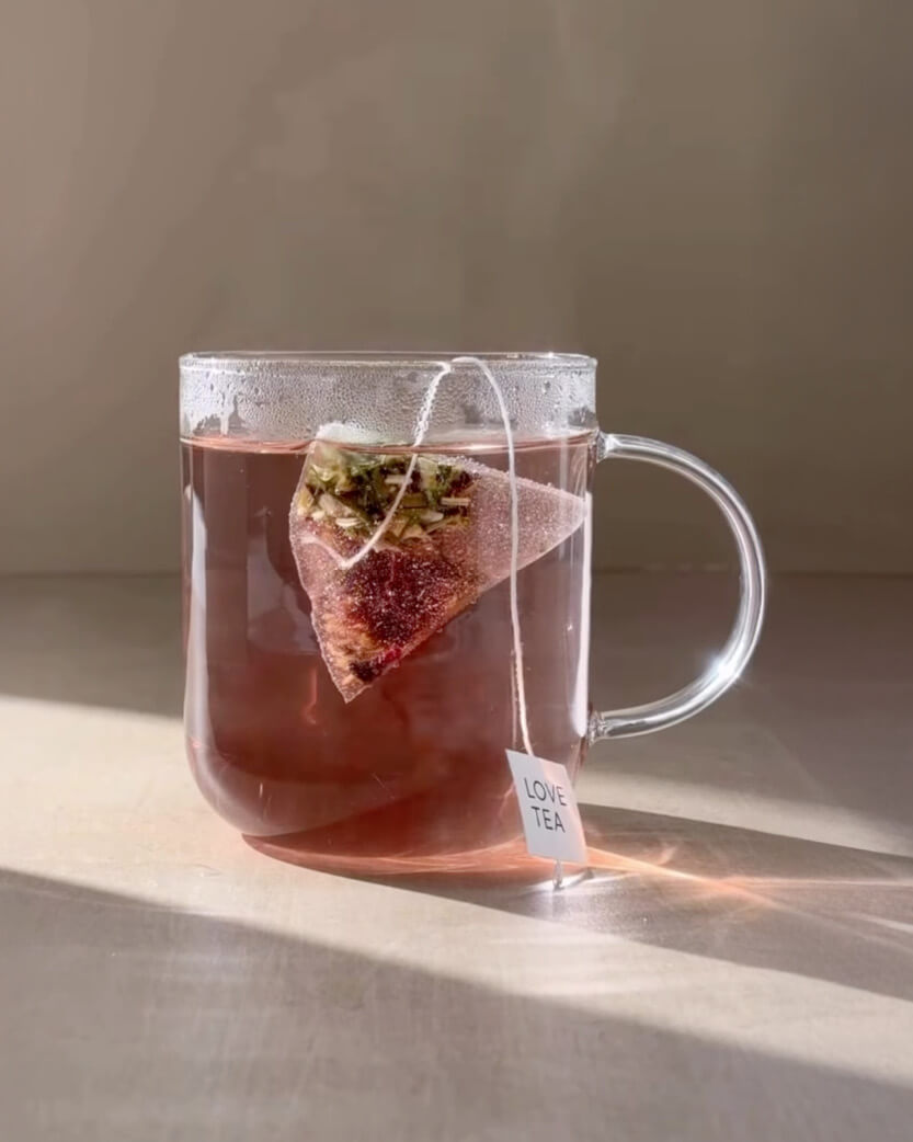 Clear mug with pink tea and a pyramid tea bag on a neutral background.