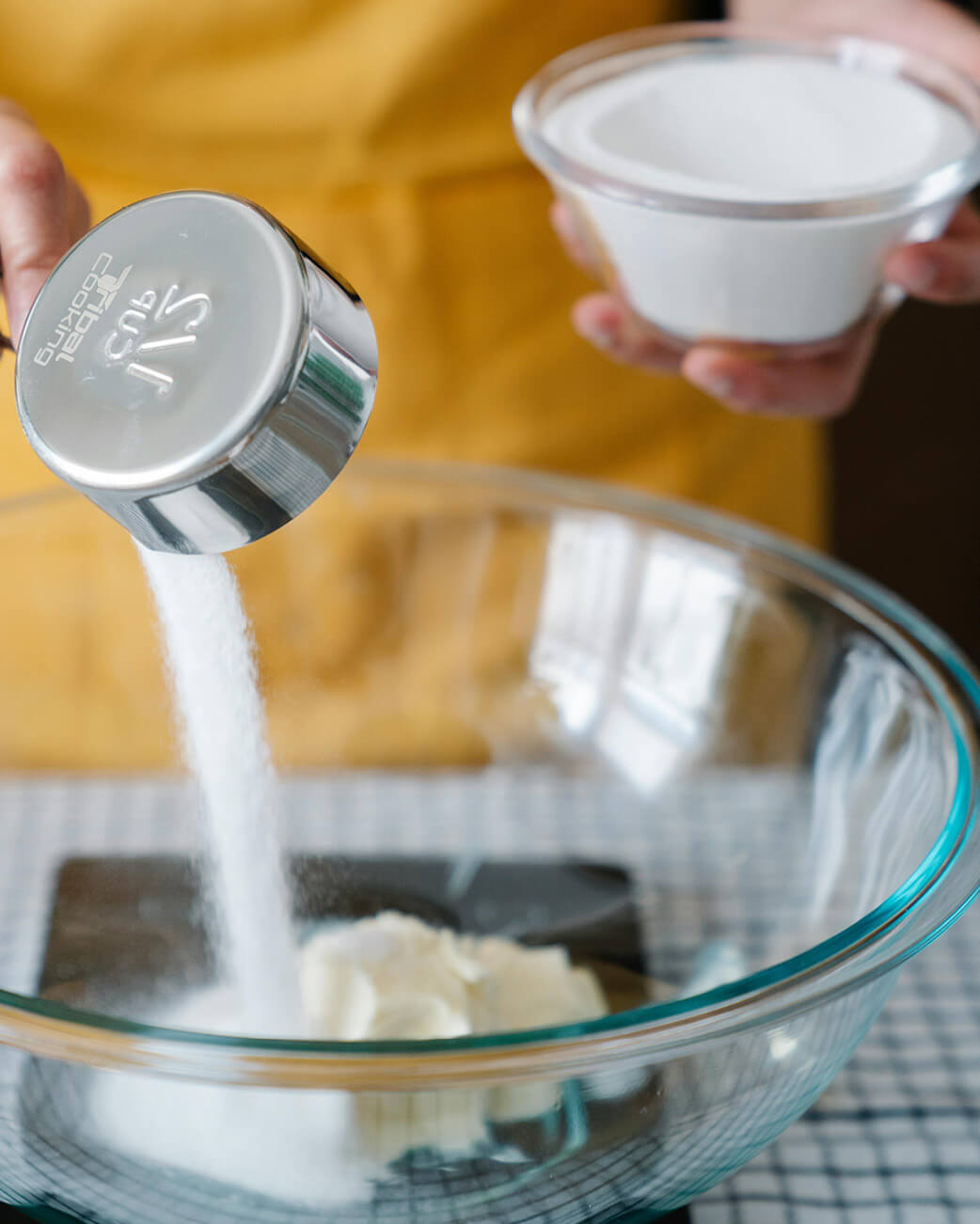Person pouring sugar from a measuring spoon into a glass bowl with a blurred background.