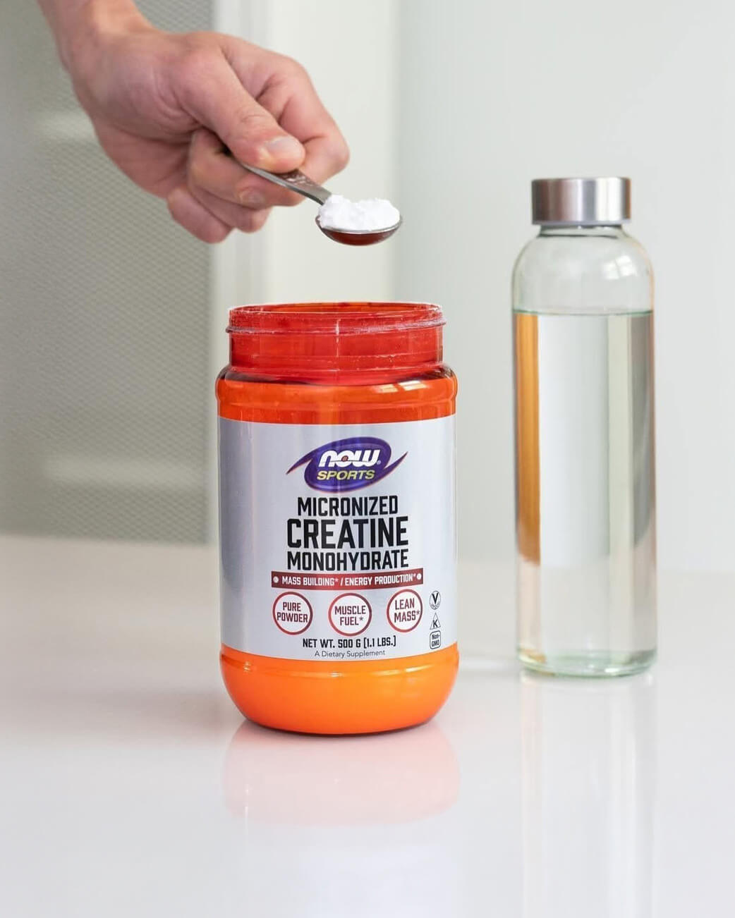 Person measuring creatine powder from a NOW Sports Nutrition container with a spoon, next to a glass of water.