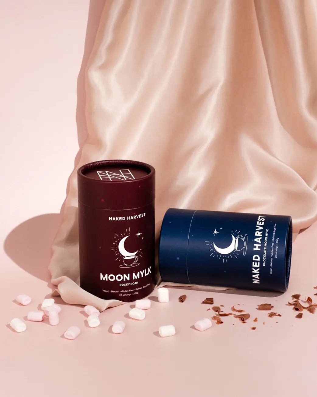 Two cylindrical containers labeled 'Naked Harvest' on a soft pink fabric background.