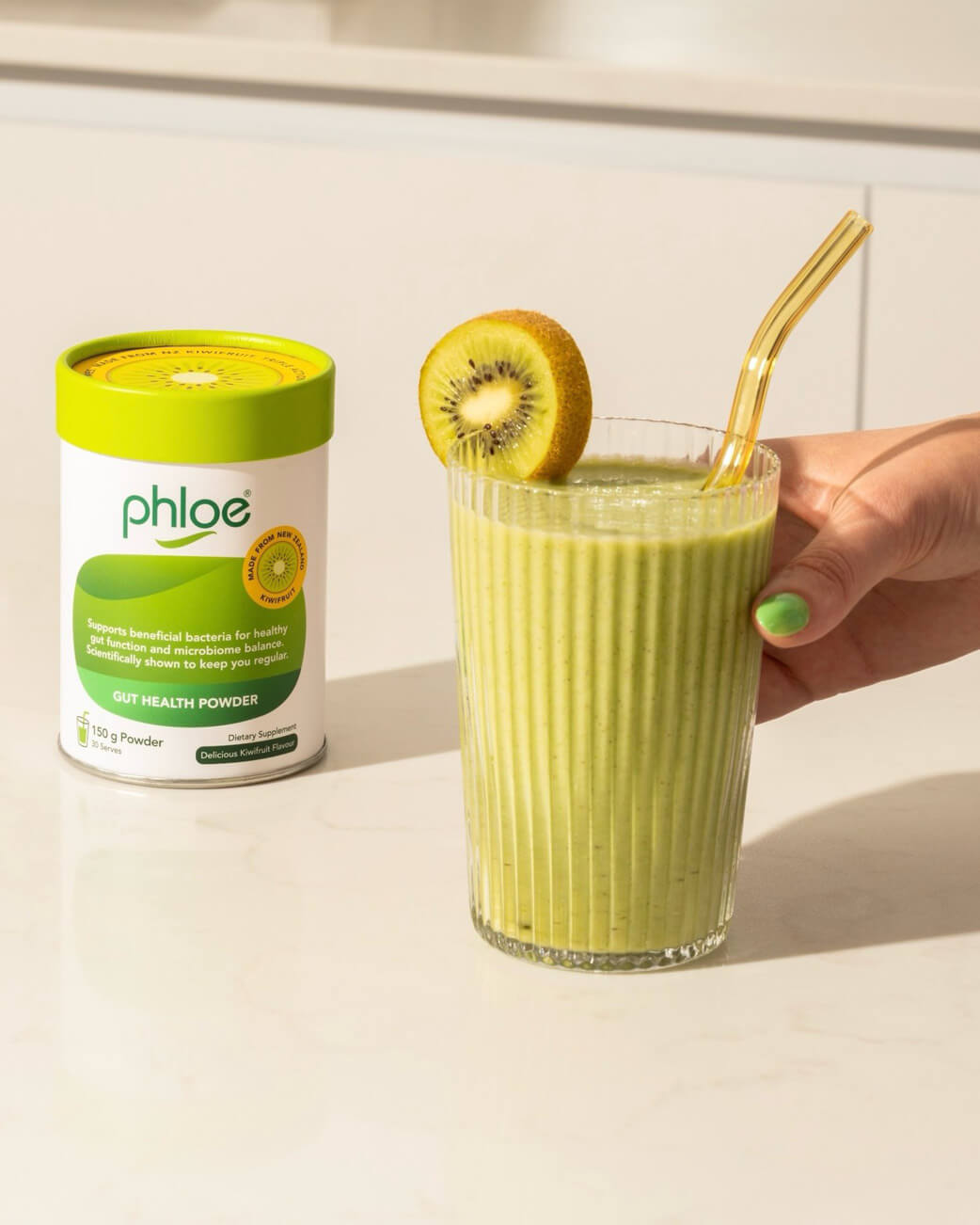 Green smoothie with a kiwi slice and gold straw next to a container of Phloe Gut Health Powder on a light background.