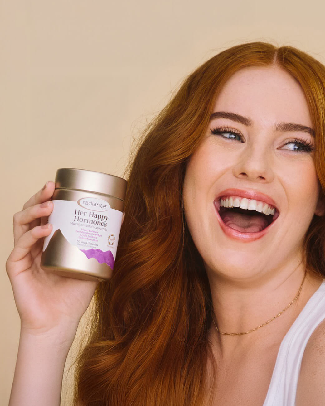 Woman with red hair holding a product container against a beige background.