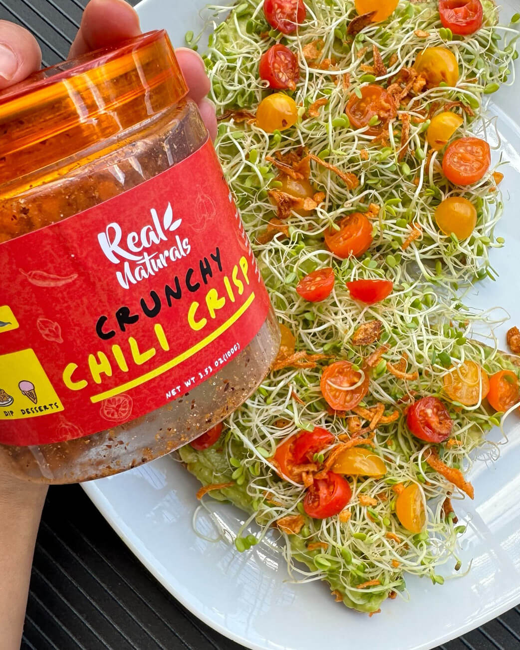 Jar of Real Nature Crunchy Chili Crisp next to a salad with cherry tomatoes on a white plate.