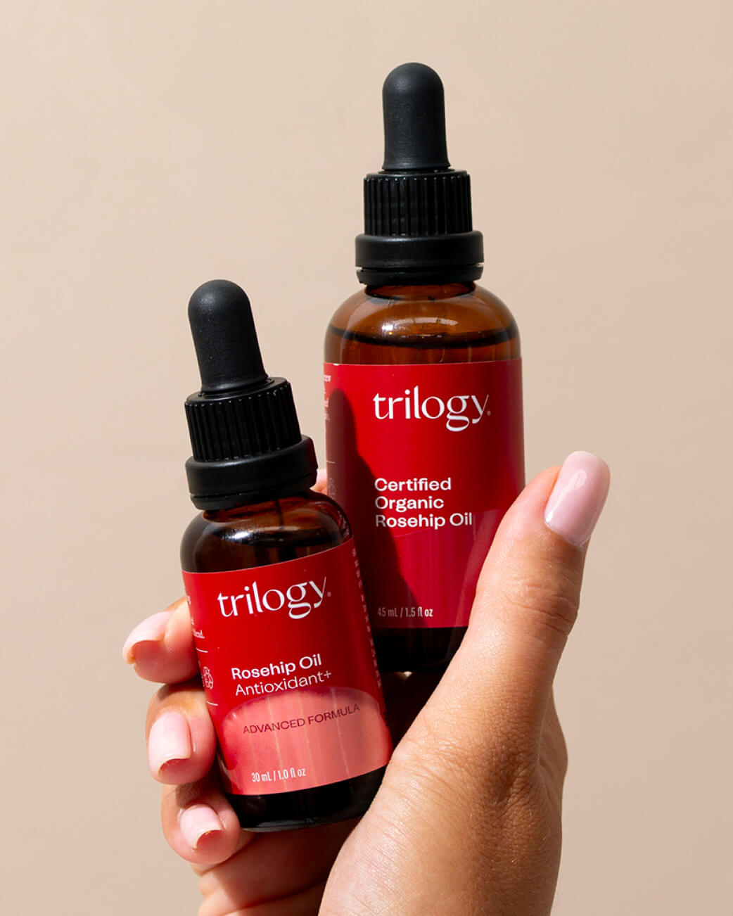 Two bottles of Trilogy rosehip oil held by a hand against a beige background.