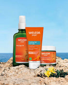 Weleda arnica products including massage oil, cooling gel, and body butter on a rocky surface with a blue sky background.