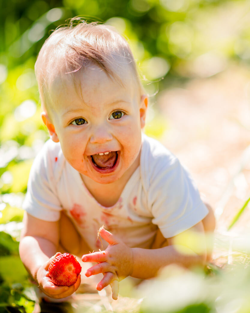 Child outdoors with a strawberry, surrounded by greenery.