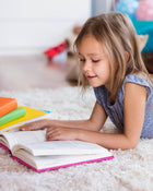Young girl reading a book on a carpeted floor with colourful books around her.