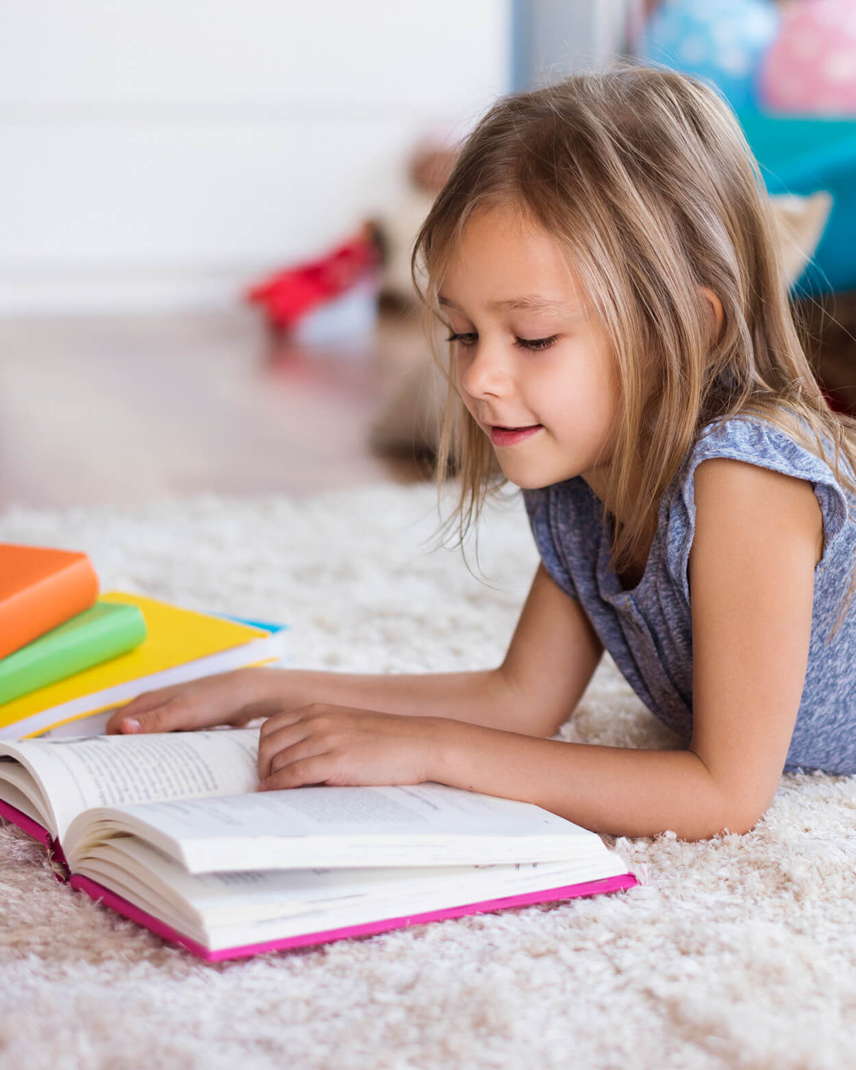Young girl reading a book on a carpeted floor with colourful books around her.
