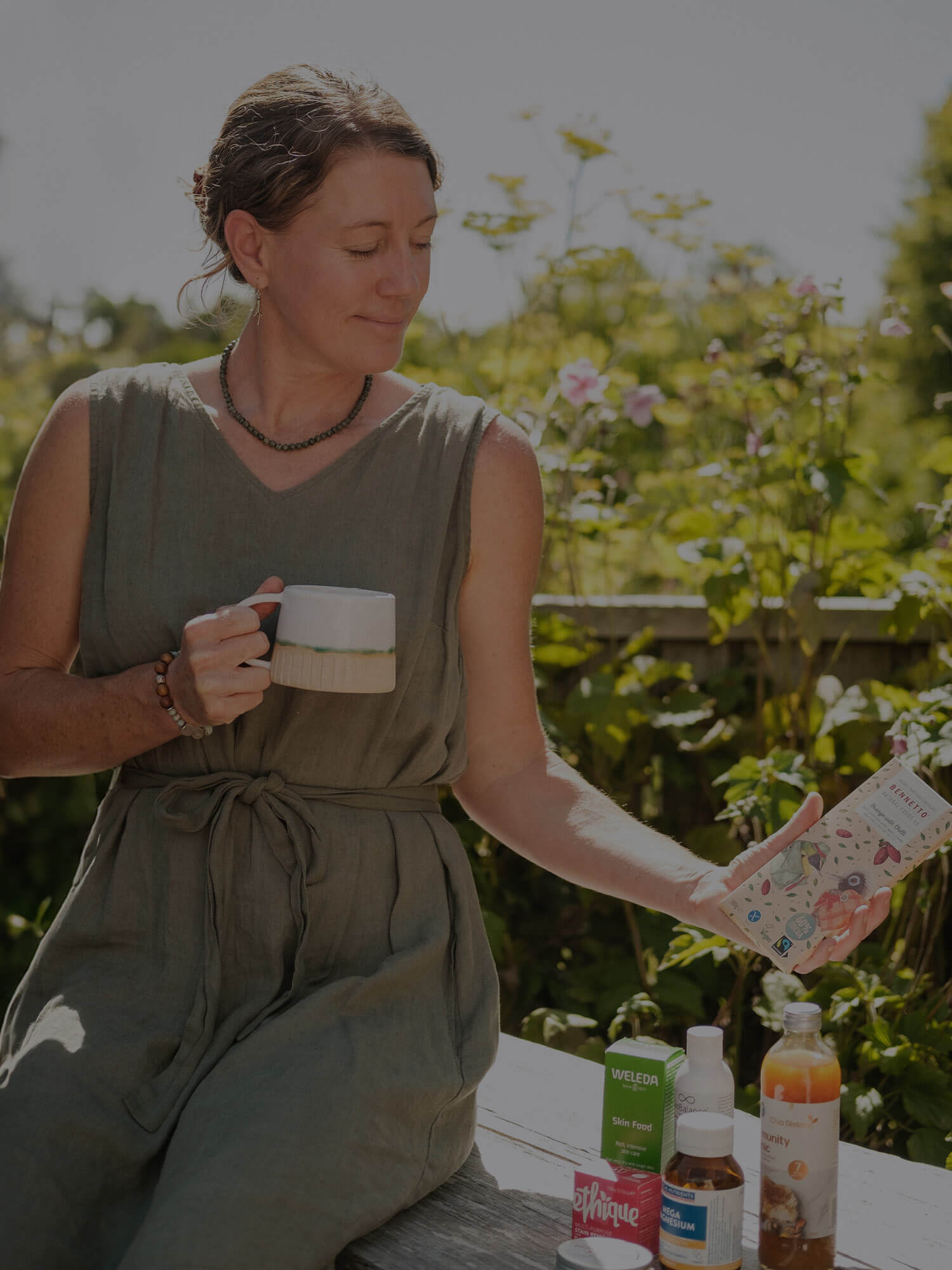 Woman sitting outdoors with a cup, chocolate, and various wellness items on a bench.