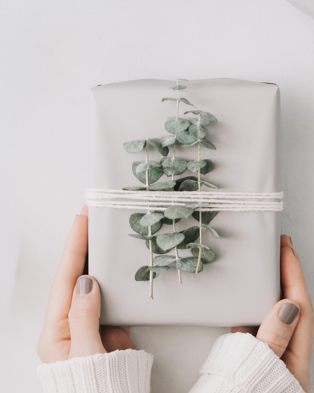 Gift wrapped in white paper with eucalyptus leaves, held by hands against a light background.