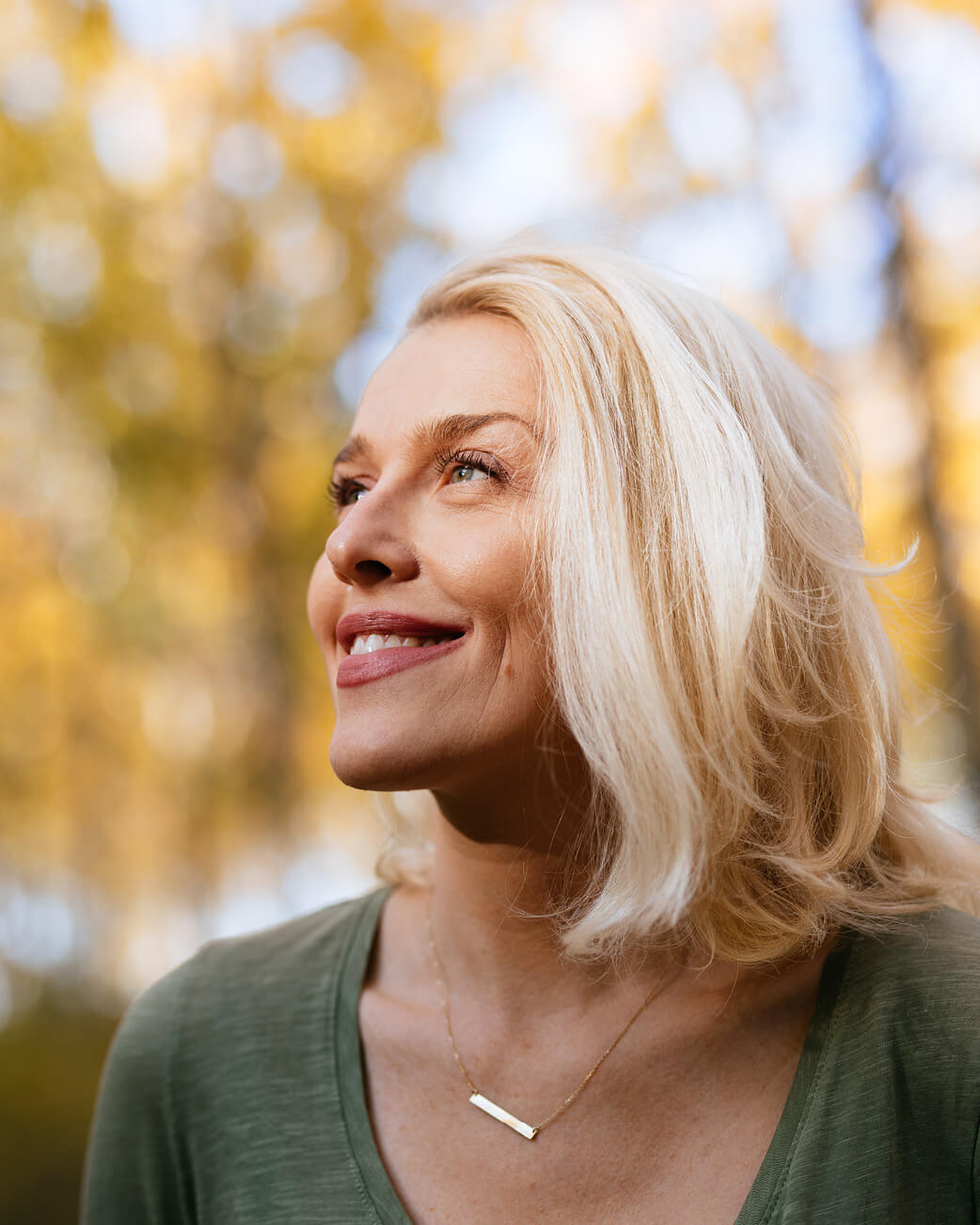 Woman with blonde hair and a green shirt standing in an outdoor setting with blurred trees in the background.