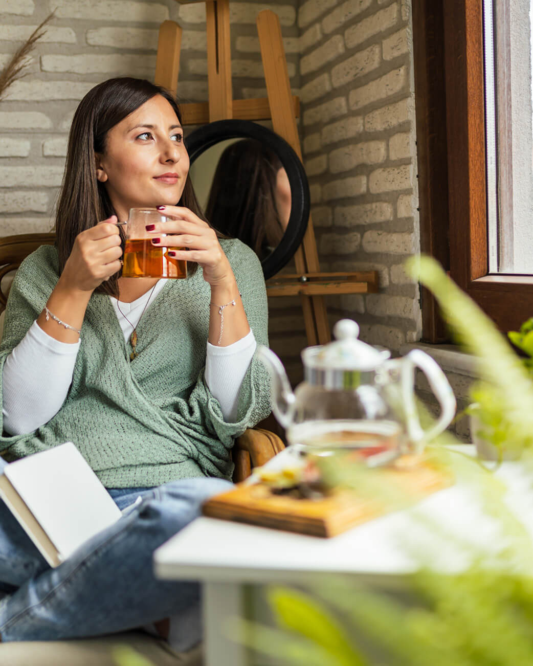 Woman sitting indoors holding a mug of tea, with a table in front of her featuring a teapot and books.