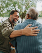 Two men embracing outdoors with trees and a lake in the background.