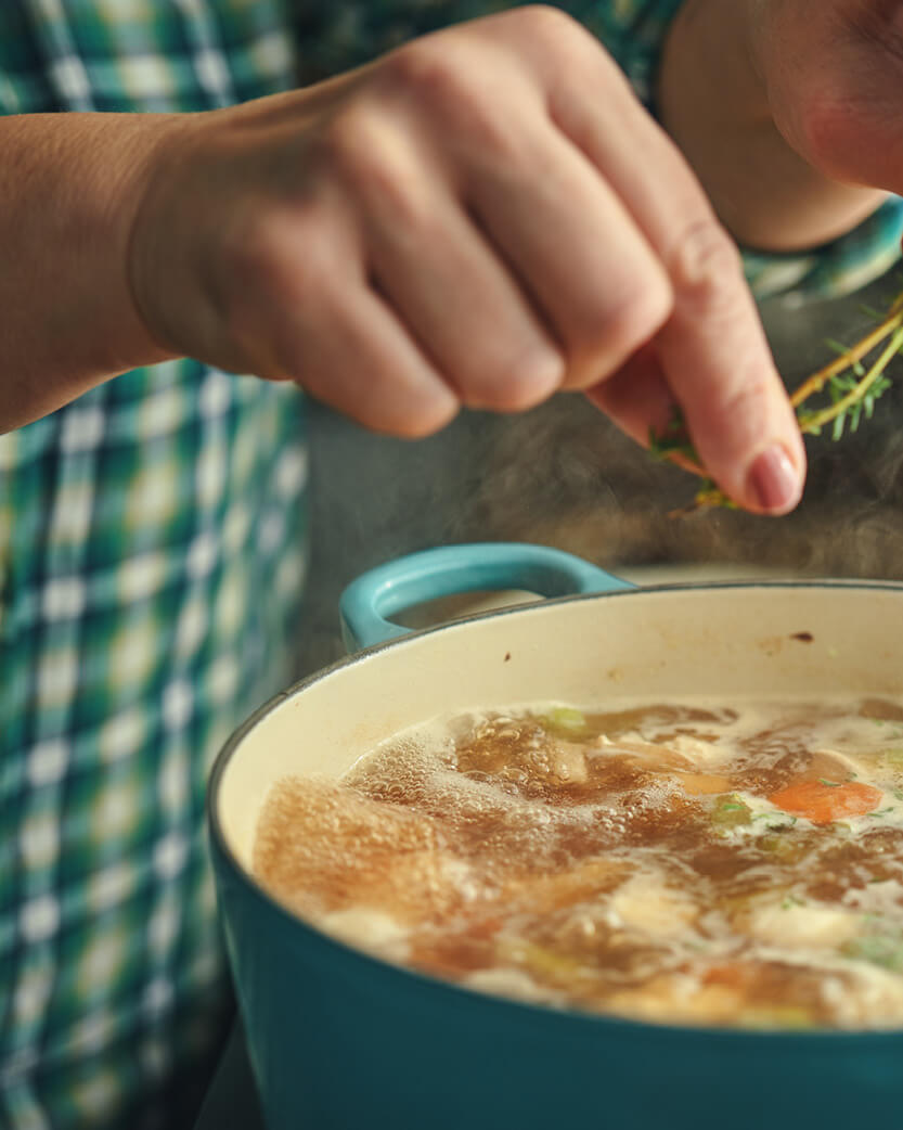 Person seasoning soup in a pot with visible vegetables
