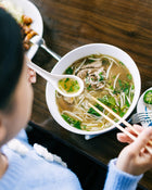 Person eating noodles with chopsticks from a bowl on a wooden table