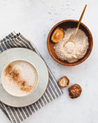 Cappuccino in a white cup with a wooden bowl of brown sugar on a light surface with mushrooms.