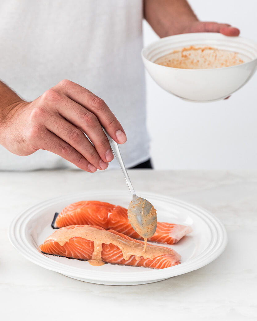 Person seasoning salmon fillets with a sauce mixture on a white plate.