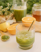 Green smoothie in a glass with a spoon, surrounded by oranges, lemons, and herbs on a light surface.